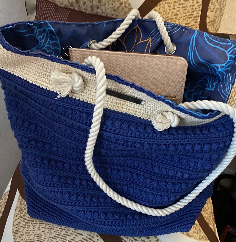 Puede incluir: Un bolso tote azul y crema con asa de cuerda. El bolso tiene un patr&oacute;n de punto texturizado y un interior floral azul. Una cartera beige es visible dentro del bolso.