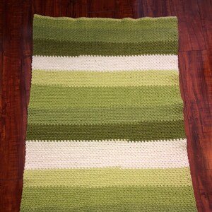 Może przedstawiać: Ręcznie robiony, prostokątny koc z poziomymi pasami w r&oacute;żnych odcieniach zieleni i złamanej bieli. Koc wykonany jest z teksturowanego, dzianinowego materiału i jest prezentowany na ciemnej drewnianej powierzchni.