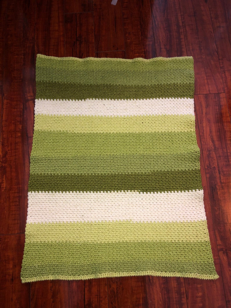 Może przedstawiać: Ręcznie robiony, prostokątny koc z poziomymi pasami w r&oacute;żnych odcieniach zieleni i złamanej bieli. Koc wykonany jest z teksturowanego, dzianinowego materiału i jest prezentowany na ciemnej drewnianej powierzchni.