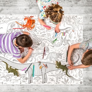 May include: Three children color a large dinosaur-themed coloring sheet with colored pencils. The sheet features a variety of dinosaurs, including a triceratops, a stegosaurus, and a brachiosaurus. The sheet is laid out on a wooden floor.
