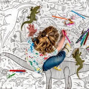May include: A child colors a large dinosaur-themed coloring sheet with colored pencils. The sheet features many different dinosaurs, including a stegosaurus, a brachiosaurus, and a triceratops. There are also several plastic dinosaur toys on the sheet.