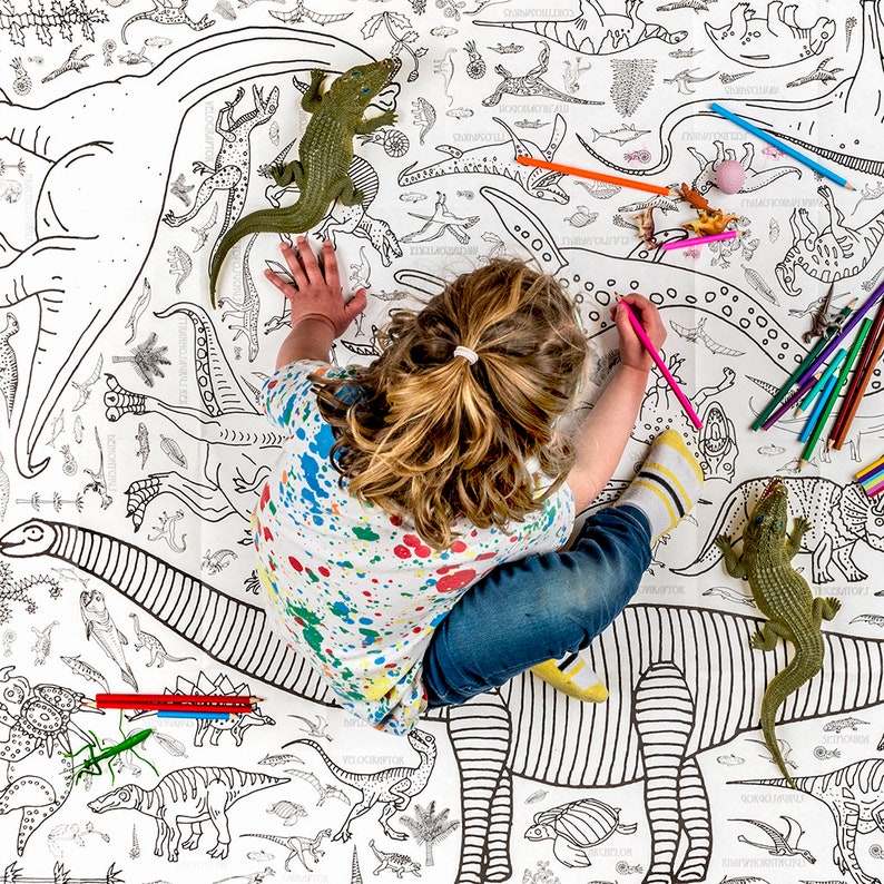 May include: A child colors a large dinosaur-themed coloring sheet with colored pencils. The sheet features many different dinosaurs, including a stegosaurus, a brachiosaurus, and a triceratops. There are also several plastic dinosaur toys on the sheet.