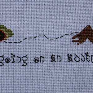 May include: Cross-stitch embroidery with a green and brown house and a mountain, connected by a dotted line. The text "I'm going on an adventure" is stitched below.