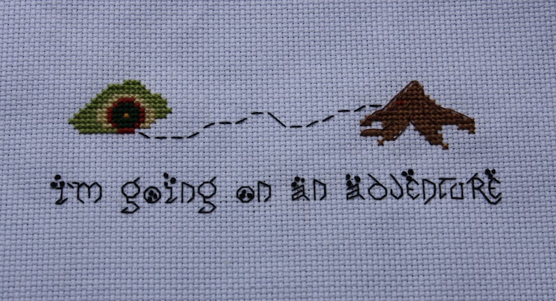 May include: Cross-stitch embroidery with a green and brown house and a mountain, connected by a dotted line. The text "I'm going on an adventure" is stitched below.