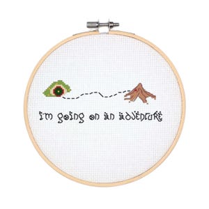 May include: A white embroidery hoop with a cross-stitch design of a green eye with a red pupil, a dotted line, and a brown mountain. The text "I'm going on an adventure" is stitched below the design.