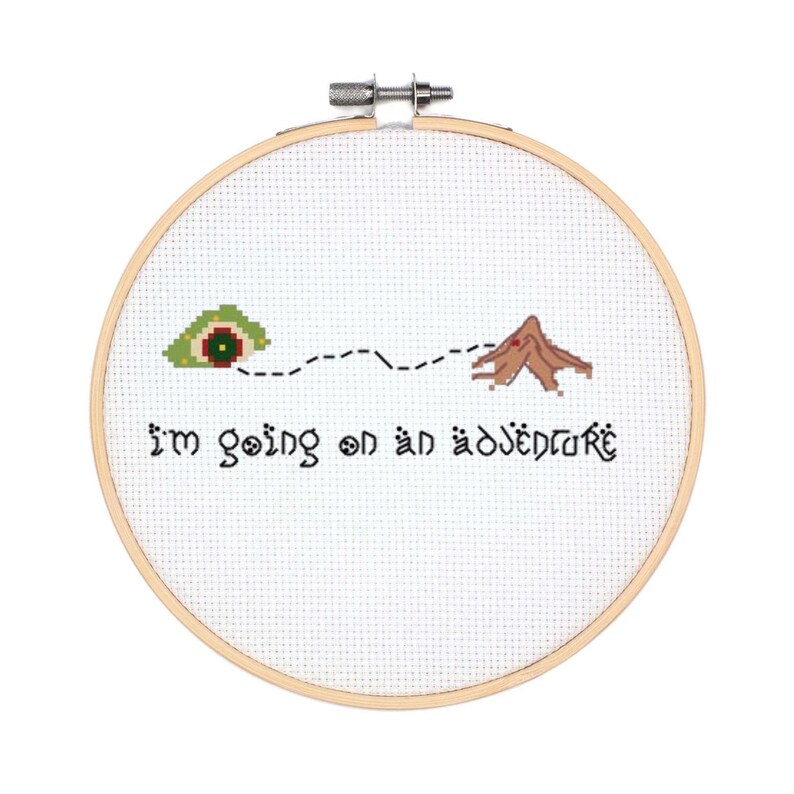 May include: A white embroidery hoop with a cross-stitch design of a green eye with a red pupil, a dotted line, and a brown mountain. The text "I'm going on an adventure" is stitched below the design.