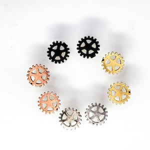 May include: An assortment of small, gear-shaped metal components in various colors, including black, copper, gold, and silver. The gears are arranged in a circular pattern against a white background, showcasing their intricate designs.