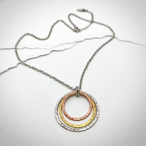 May include: A silver chain necklace with a pendant featuring three concentric circles. The outer circle is silver, the middle circle is gold, and the inner circle is copper. The circles have a textured finish.