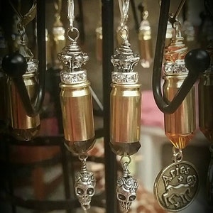 Puede incluir: Un par de pendientes de casquillo de bala dorado con un dije de calavera de plata y un dije de corona de plata. Los pendientes cuelgan de un expositor negro.