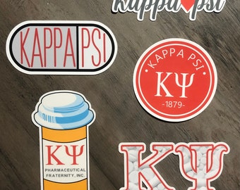 May include: A collection of stickers featuring the Greek letters and name of Kappa Psi Pharmaceutical Fraternity, Inc. Designs include a pill capsule, a prescription bottle, and a circular emblem, all in red and white. The stickers are on a wooden surface.