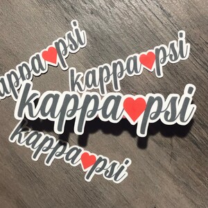 May include: White die-cut stickers with the words "kappa psi" in a script font, with a red heart replacing the letter "a". The stickers are scattered on a wooden surface.