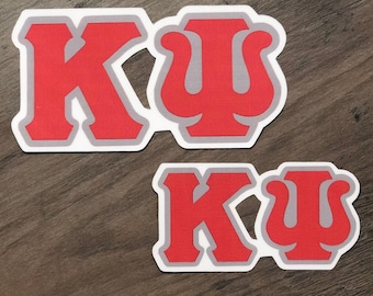 May include: Two red and gray stickers with the Greek letters Kappa and Psi. The letters are outlined in gray and have a white border. The stickers are on a wooden surface.