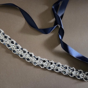 May include: A navy blue bridal belt with a sparkling, ornate design. The belt features a row of clear and dark blue crystals, and is attached to a long, navy blue satin ribbon.