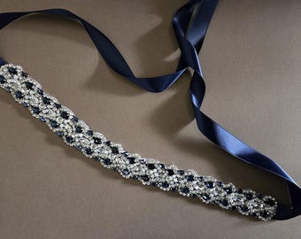 Op de afbeelding: Een marineblauwe bruidsriem met een sprankelend, sierlijk ontwerp. De riem heeft een rij heldere en donkerblauwe kristallen en is bevestigd aan een lang, marineblauw satijnen lint.