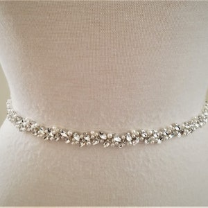 May include: A delicate bridal sash with a beaded design featuring clear rhinestones and faux pearls. The sash is white and has a satin finish.