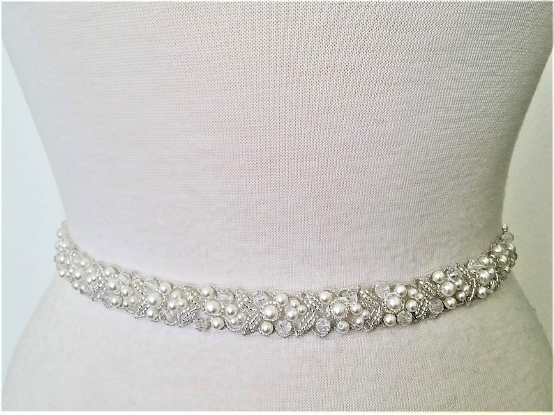 May include: A white beaded bridal sash with pearl and crystal embellishments. The sash is designed to be worn around the waist.