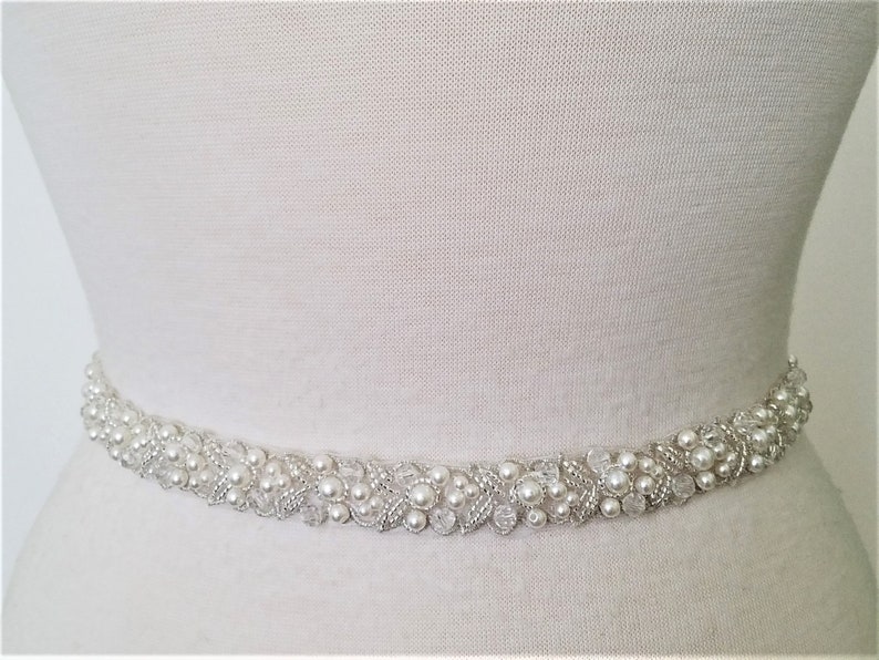 May include: A white beaded bridal sash with pearl accents. The sash is made of a delicate fabric and is adorned with small, white pearls and silver beads.
