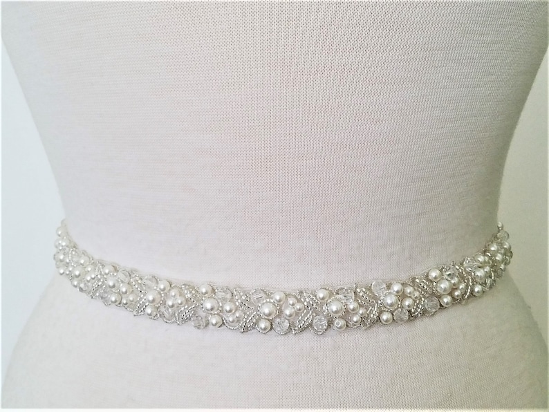 May include: A white beaded bridal belt with pearl and crystal embellishments. The belt is designed to be worn around the waist.