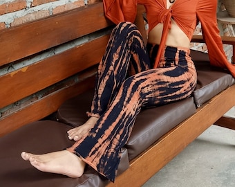 May include: A woman wearing a rust-coloured, long-sleeved top with a tie front and black and orange tie-dye flared trousers. She is sitting on a wooden bench with a brown leather cushion.