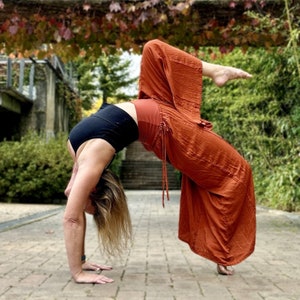 May include: A person in a yoga pose, arched backward with hands on the ground. They are wearing a black top and rust-colored pants. The background includes a brick walkway, greenery, and a bridge.