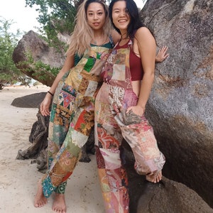 Può includere: Due persone indossano salopette in patchwork, in piedi davanti a una grande roccia. Le salopette sono fatte di diversi tessuti e colori, tra cui marrone, verde, rosso e blu. Le persone sorridono e guardano la fotocamera.