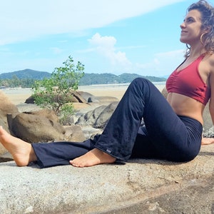 May include: A woman wearing a red sports bra and black yoga pants sits on a rock with her legs crossed. She is looking off to the side and has her hair pulled back. The background is a beach with rocks and water.