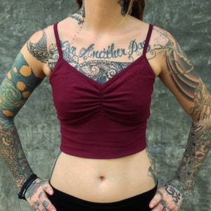 Op de afbeelding: Een diepbordeauxrode crop top met een V-hals, een geplooid detail in het midden en dunne schouderbandjes. Het kledingstuk is effen donkerrood van kleur en heeft een nauwsluitend model dat boven de taillelijn eindigt voor een strak ontwerp.