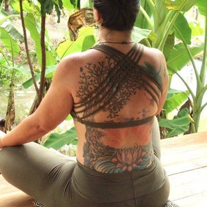 May include: A woman sits cross-legged on a wooden deck with a green leafy background. She is wearing a green yoga top with a criss-cross back and a large tree tattoo on her back. She is also wearing green yoga pants.