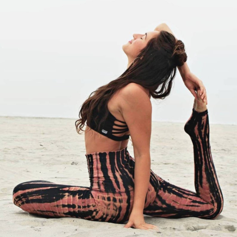May include: A woman in a black sports bra and tie-dye leggings performs a yoga pose on a sandy beach. Her hair is long and dark, and she is looking up at the sky.
