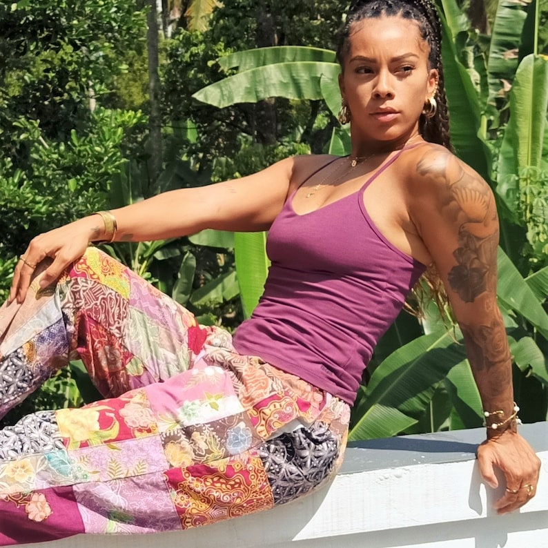May include: A woman wearing a purple tank top and colorful patchwork pants sits on a white surface. She has a tattoo on her arm and is wearing gold jewelry.