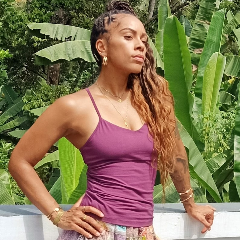 May include: A woman with long, wavy brown hair wearing a purple tank top and gold jewelry. She is standing in front of a lush green background.