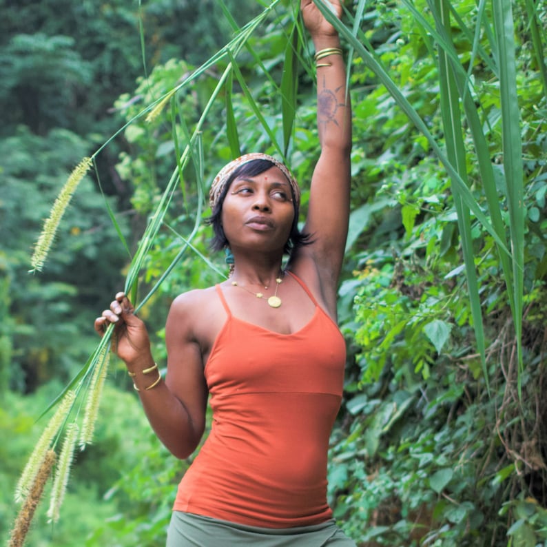 May include: A woman with a patterned headband and a red tank top stands in a lush green forest. She is holding a long stalk of grass with her left hand and her right arm is raised above her head.