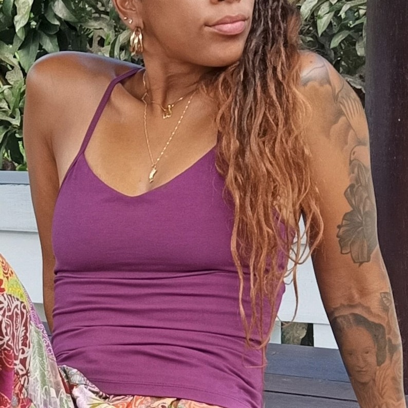 May include: A woman wearing a purple tank top with a gold necklace and hoop earrings. She has long, curly brown hair and a tattoo on her arm.