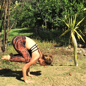 May include: A woman performs a yoga pose outdoors, balancing on her hands with her legs raised in the air. She is wearing a black sports bra with a criss-cross design and orange and brown tie-dye leggings.