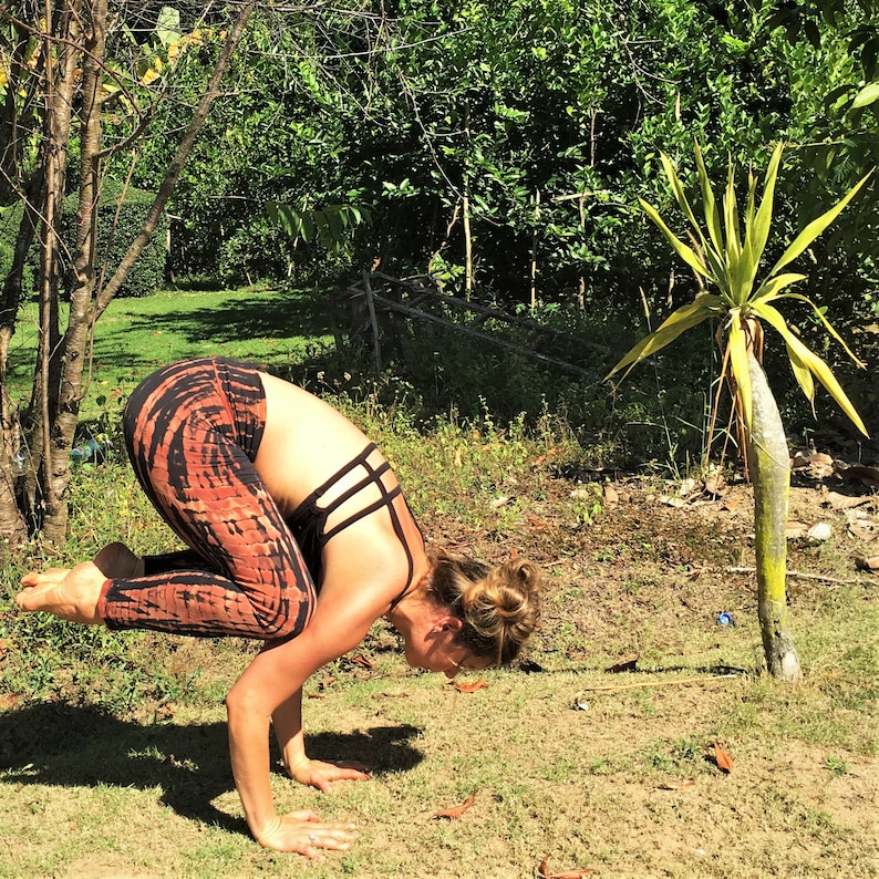 May include: A woman performs a yoga pose outdoors, balancing on her hands with her legs raised in the air. She is wearing a black sports bra with a criss-cross design and orange and brown tie-dye leggings.