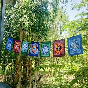 May include: Seven colorful fabric flags with mandala designs hang from a tree branch. The flags are blue, green, red, orange, and purple. The flags are decorated with intricate patterns and symbols.