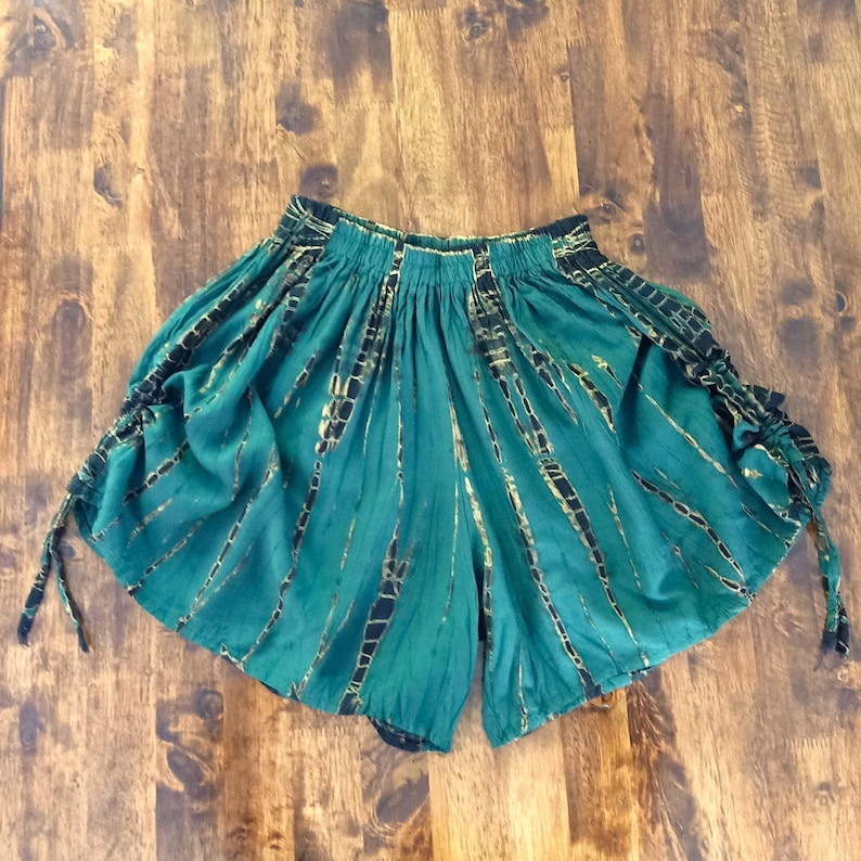 May include: Teal-colored shorts with a tie-dye pattern featuring black and gold accents. The shorts have an elastic waistband and drawstring ties on the sides. The fabric has a flowing, lightweight appearance.