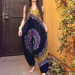 Op de afbeelding: Een mouwloze jumpsuit met een tie-dye patroon met een groot spiraalontwerp in tinten paars, zwart en geel. Het kledingstuk heeft een diepe V-hals, een relaxte pasvorm rond de romp en losse, toelopende broekspijpen die bij de enkels samenkomen.