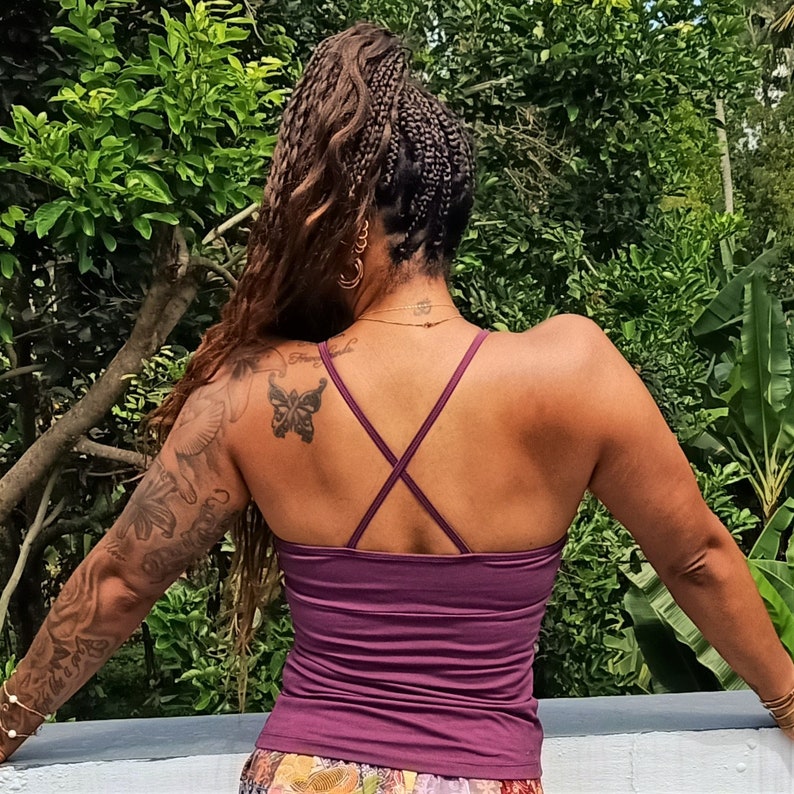 May include: A woman with long, braided hair is wearing a purple tank top with a criss-cross back. She has a butterfly tattoo on her shoulder.