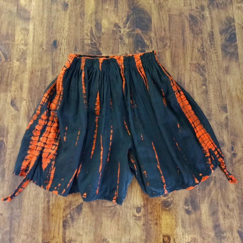 May include: Black and orange tie-dye shorts with an elastic waistband. The shorts have a loose, flowing design with orange vertical stripes against a black background. The shorts are made of a lightweight fabric.