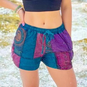 May include: Patchwork shorts featuring a vibrant mix of teal, purple, and magenta fabric panels with intricate circular patterns and sun motifs. These shorts include an elastic waistband with a matching drawstring tie for a secure fit.