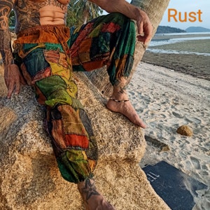 May include: Patchwork pants in shades of green, orange, and brown. The pants have an elastic waistband and cuffs. The image shows the pants on a person sitting on a rock near a beach. The word "Rust" is in the top right corner.