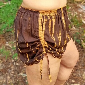 May include: Brown and gold tie-dye shorts with a gathered drawstring waist. The shorts have a loose, comfortable fit and a unique pattern. The fabric appears to be lightweight and suitable for warm weather.