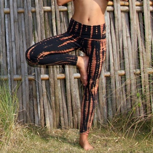 May include: Black and orange tie-dye leggings with a high waist and a wide waistband. The leggings are worn by a person standing in front of a bamboo fence.