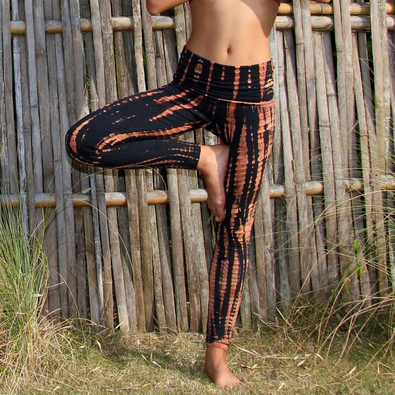 May include: Black and orange tie-dye leggings with a high waist and a wide waistband. The leggings are worn by a person standing in front of a bamboo fence.
