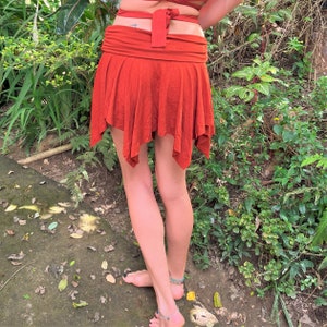 May include: A rust-colored, asymmetrical skirt with a wide waistband. The skirt is made of a soft, flowing fabric and has a relaxed fit.