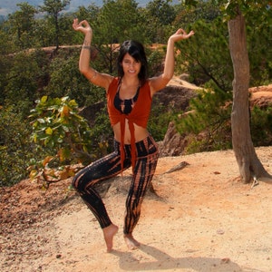 May include: A woman wearing a rust-colored crop top with a tie front and black and brown tie-dye leggings poses with her arms raised in a yoga pose.