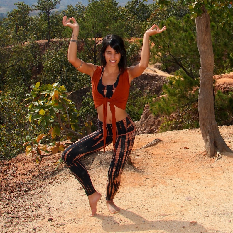 May include: A woman wearing a rust-colored crop top with a tie front and black and brown tie-dye leggings poses with her arms raised in a yoga pose.