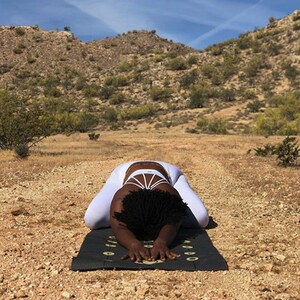 Pu&ograve; includere: Una persona con un abbigliamento da yoga bianco pratica yoga su un tappetino da yoga nero con segni bianchi in un paesaggio desertico.