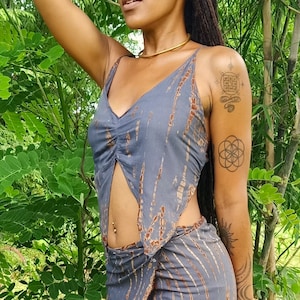 May include: A grey and brown tie-dye crop top with a low-cut neckline and a slit at the front. The top is worn with a matching grey and brown skirt. The model is wearing a gold choker necklace and has several tattoos on her arm.